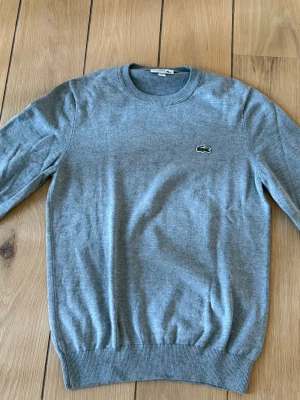 Grå stickad tröja från Lacoste - Säljer en stilren grå stickad tröja från Lacoste med rund halsringning och det klassiska krokodilmärket broderat på bröstet. Tröjan har långa ärmar och ribbade muddar vid ärmslut och nederkant. Perfekt för lager på lager och enkel att matcha.