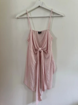 Ljusrosa linne M - Ljusrosa linne med smala axelband och djup v-ringning. Klänningen har ett långt knytband framtill som ger en snygg detalj och en avslappnad siluett. Perfekt för sommardagar och enkel att styla.