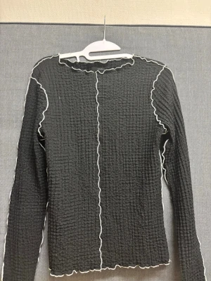 Svart långärmad topp med vågiga kanter - Trendig svart långärmad topp från Monki med vågiga kontrastsömmar i vitt längs kanter och ärmar. Toppen har en ribbad, strukturerad yta och figurnära passform. Perfekt att styla med jeans eller kjol för en cool look.