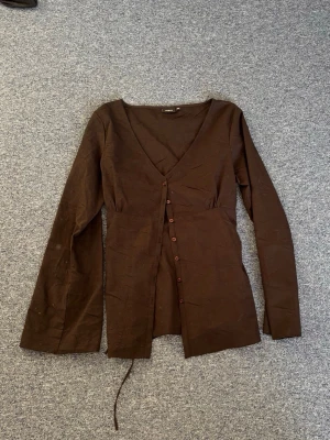 Svart blus med knappar och snörning  - Snygg svart blus från Lindex med långa ärmar och v-ringning. Blusen har knappar framtill och ett knytband i midjan som ger en figurnära passform. Tillverkad i ett lätt tyg som känns skönt mot huden. Perfekt att styla med jeans eller kjol.  Oanvänd och utan defekter, fläckarna är endast damm då den aldrig kommit till användning. 