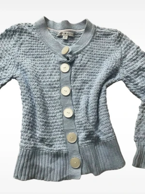 Coquette Ljusblå stickad kofta med stora knappar Tommy Hilfiger  - Snygg ljusblå stickad kofta från Tommy Hilfiger med stora vita knappar framtill. Koftan har ribbade muddar vid ärmslut och nederkant samt en rundad krage. Materialet är mjukt och strukturerat, perfekt för lager på lager.