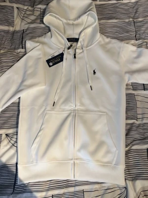Vit hoodie från Polo Ralph Lauren - Snygg vit hoodie från Polo Ralph Lauren med dragkedja framtill, två stora fickor och klassisk huva med dragsko. Ikoniska svarta loggan broderad på bröstet. Tillverkad i mjukt material som känns skönt mot huden. Perfekt för en clean och stilren look.