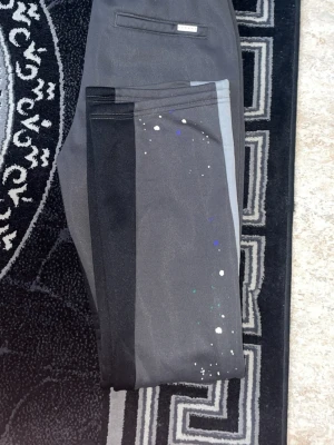 Grå-svarta mjukisbyxor med färgstänk - Säljer ett par grå och svarta mjukisbyxor med coola färgstänk i vitt, blått och grönt. Byxorna har en rak passform och en ficka bak. Perfekta för en avslappnad och trendig look.