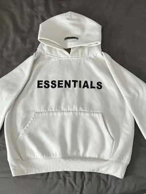 Vit Essentials hoodie med tryck - Snygg vit hoodie från Essentials med svart texttryck på bröstet. Klassisk känguruficka framtill, ribbade muddar och stor huva. Använt fåtal gånger. Pris kan diskuteras. 