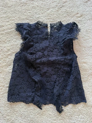 Svart spetsblus från Zara - Säljer en marinblå spetsblus från Zara med vackra spetsdetaljer och ärmlös design. Blusen har en liten ståkrage och knäppning i nacken. Perfekt för dig som gillar en elegant och trendig look. Knappt använd. 