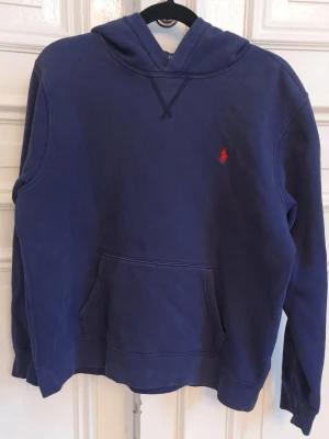 Marinblå hoodie från Polo Ralph Lauren - Snygg marinblå hoodie från Polo Ralph Lauren med klassisk röd logga broderad på bröstet. Hoodien har huva, känguruficka och ribbade muddar. Junior storlek 13-14 år. Kommer från ett djur och rökfritt hem. 