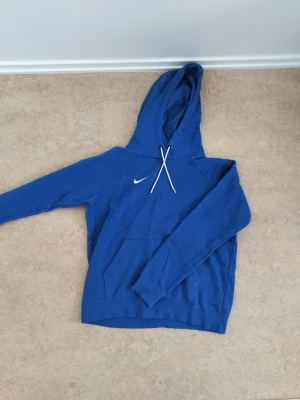 Blå Nike hoodie med luva - Snygg blå hoodie från Nike med klassisk vit logga på bröstet och matchande vita dragsnören i huvan. Hoodien har en stor magficka och ribbade muddar vid ärmslut och nederkant. Perfekt för en avslappnad och sportig stil. Polyster