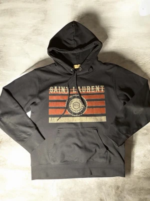 Svart hoodie från Saint Laurent - Svart hoodie från Saint Laurent med huva och dragsko. Framsidan har ett tryck med röda och vita ränder samt ett emblem och texten 'SAINT LAURENT'. Känguruficka framtill och ribbade muddar. Perfekt för en cool och avslappnad stil.