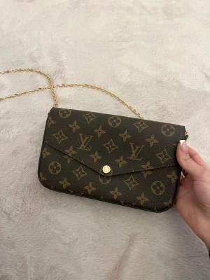 Brun handväska - Snygg brun handväska med klassiskt monogrammönster i guld och beige. Väskan har en guldfärgad kedjerem och knappstängning framtill. 