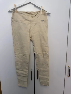 Vita leggings med strukturmönster - Säljer ett par beige/krämvita leggings med hög midja och snyggt strukturmönster. De är stretchiga och har en diskret logga framtill i midjan. Perfekta för dig som gillar en enkel men trendig look. Materialet känns mjukt och följsamt.