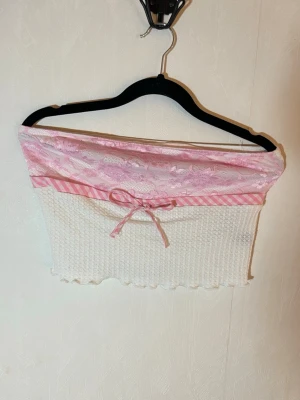 Söt vit och rosa tubtopp med spets - Tubtopp i vitt med ljusrosa spets upptill och rosa/vit-randigt band med rosett framtill. Toppen är croppad och har en vågig kant nertill. Perfekt för dig som gillar gulliga detaljer och pastellfärger. Helt ny, aldrig använd. Shein, storlek M. Djur, rök och parfymfritt hem.