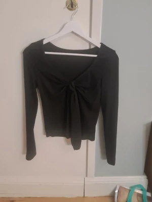 Svart långärmad topp med knyt - Snygg svart långärmad topp med knyt framtill och djupare ringning. Modellen är figurnära och croppad, vilket ger en trendig look. Perfekt att matcha med höga jeans eller kjol. Materialet känns mjukt och stretchigt.
