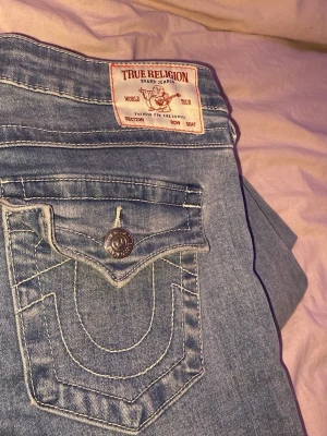 True Religion ljusblå jeans - Ljusblå jeans från True Religion med klassisk kontrastsöm och snygga detaljer på bakfickan. Jeansen har en unik fickdesign med silverfärgade knappar och patch med logotyp på baksidan. Materialet är denim och passformen är bootcut 