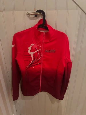 Röd All Star Gymnastics jacka - Ord pris ca 600:- Snygg rosa (ej röd) träningsjacka från All Star Gymnastics med hel dragkedja, hög krage och vita detaljer. Framsidan har ett gymnastikmotiv och logga, baksidan har stor stjärna och text. Tillverkad i stretchigt funktionsmaterial, perfekt för träning eller uppvärmning.