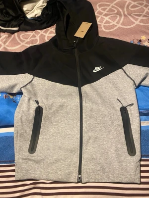  Nike tech - Nike hoodie i grått och svart med hel dragkedja framtill och två stora fickor med dragkedja. Klassisk Nike-logga på bröstet och huva med snörning. Tillverkad i mjukt sweatshirtmaterial, perfekt för en sportig och avslappnad stil.