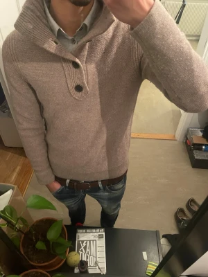 Beige stickad ull Hugo boss tröja  - Säljer en biege stickad tröja med ribbad hög krage och två knappar framtill. Tröjan har långa ärmar och är tillverkad i ett mjukt, värmande material. Perfekt för kyliga dagar och enkel att matcha med jeans eller chinos.