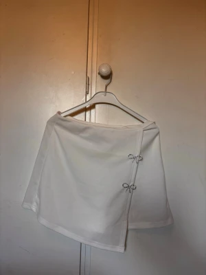 Skort vit, kjol-shorts - Ej använda supersnygg vit skort med omlottkänsla framtill och två glittriga strassrosetter som detaljer. Skorten har en clean look och är gjord i ett mjukt tyg som känns bekvämt. Perfekt för dig som vill ha kjol-looken men ändå känna dig fri att röra dig.