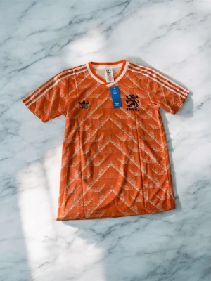 Holländsk retro fotbollströja Adidas - Orange retro fotbollströja från Adidas med vita detaljer och klassiska ränder på axlarna. Tröjan har svart Holland-lejonmärke och Adidas-logga fram, samt texten 'VAN BASTEN' och siffran 12 bak. Tillverkad i lätt och ventilerande material.