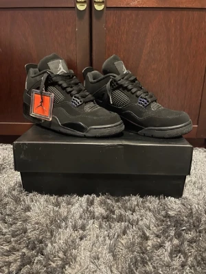 Air Jordan 4 Retro Black Cat sneakers - Snygga helsvarta Air Jordan 4 Retro Black Cat sneakers med ikonisk Jumpman-logga på plösen. Skorna har meshpaneler, mockadetaljer och robust sula. Klassisk siluett med svarta skosnören och plastdetaljer vid snörningen. Perfekta för dig som gillar streetwear och stilren design.