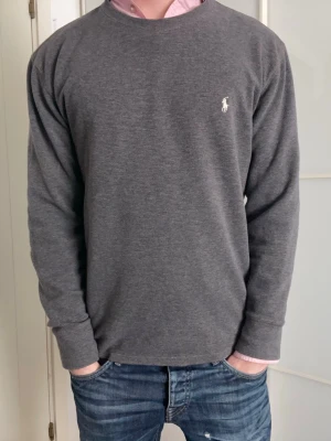 Grå långärmad tröja Polo Ralph Lauren - Snygg grå långärmad tröja från Polo Ralph Lauren med klassisk rund halsringning och vit broderad logga på bröstet. Tillverkad i mjuk bomullsmix som känns skön mot huden. Perfekt till jeans eller över en skjorta för en clean look.