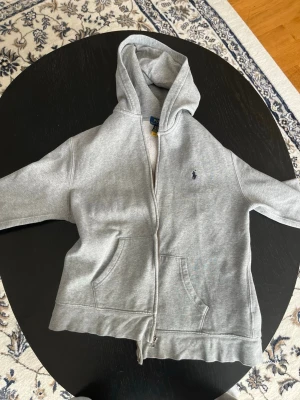 Grå zip hoodie från Polo Ralph Lauren storlek XL för barnFÖR BARN  - Klassisk grå hoodie från Polo Ralph Lauren med dragkedja framtill och två stora fickor. Hoodien har en broderad logga på bröstet och är tillverkad i mjuk bomullsblandning. Perfekt för en avslappnad och stilren look.