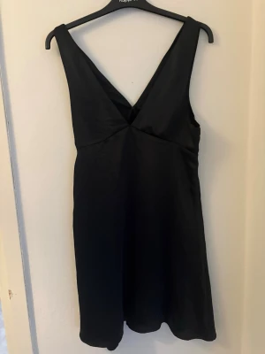 Svart omlott kortklänning - Stilren svart kortklänning med omlottdetalj och djup v-ringning både fram och bak. Klänningen har breda axelband och är ärmlös, vilket ger en elegant och enkel look. Tillverkad i ett mjukt, glansigt material som faller snyggt.