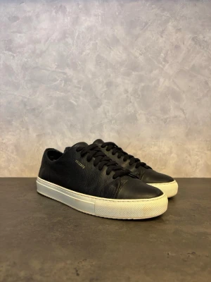 Axel Arigato Clean 90 - Stilrena svarta sneakers från Axel Arigato i mjukt läder med diskret guldfärgad logga på sidan. Skorna har klassisk låg profil, svarta skosnören och vit, tjock gummisula för en clean look. Perfekta för dig som gillar minimalistisk och modern stil. I mycket bra skick!