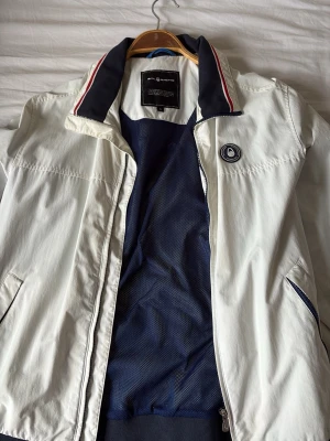 Vit bomberjacka från Alpha Industries - Snygg vit bomberjacka från Sail Racing med marinblå detaljer och röd-vit rand på kragen. Jackan har två sidofickor med dragkedja, ribbade muddar och meshfoder. Patch med amerikansk flagga på ärmen och logotyp på bröstet. Perfekt för en sportig streetwear-look.