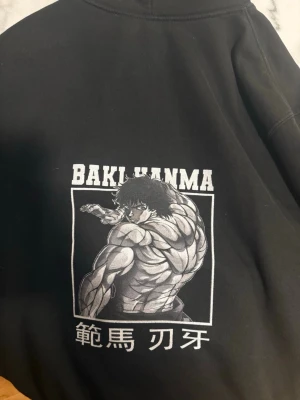 Svart gymtröja Baki Hanma hoodie animeprint - Cool svart hoodie med stort animeprint av Baki Hanma på ryggen och japansk text. Hoodien har klassisk passform, huva och är tillverkad i mjukt bomullsmaterial. Perfekt för dig som gillar manga och streetwear. Den är liten i storlek så den är mer som M