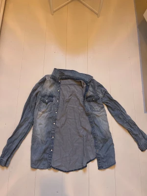 Ljusblå jeansjacka med tryckknappar - Snygg ljusblå jeansjacka med klassisk krage och tryckknappar framtill. Två bröstfickor med lock och knappdetaljer ger en cool vintagekänsla. Perfekt oversized passform och mjukt denimtyg som är lätt att styla till olika outfits.