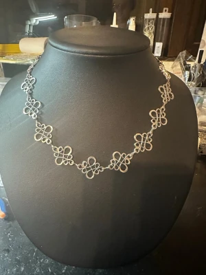 Silvrigt halsband med blommiga länkar - Unikt halsband i silverfärgad metall med dekorativa blommiga länkar som ger en elegant och lekfull look. Halsbandet består av flera sammankopplade blommönster i tunn metalltråd, vilket gör det lätt och smidigt att bära. Perfekt för dig som gillar detaljerade accessoarer.