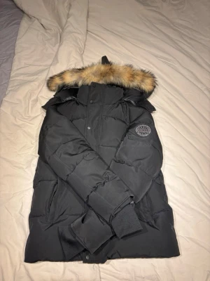 Svart dunjacka från Canada Goose - Snygg svart dunjacka från Canada Goose med klassisk patch på ärmen och detaljerade knappar. Jackan är fodrad och har en stilren design som håller dig varm under kalla vinterdagar.