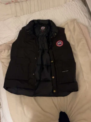 Svart dunväst från Canada Goose - Säljer en svart dunväst från Canada Goose med klassisk logga på bröstet och tryckknappar framtill. Västen har hög krage, två stora fickor och är fodrad med dun för extra värme. Perfekt för lager på lager och riktigt snygg streetstil.