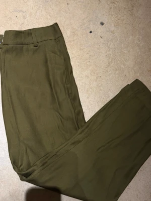 Olivgröna kostymbyxor - Snygga olivgröna kostymbyxor med raka ben och klassiska bälteshällor. Perfekta för en clean och stilren look. Materialet känns mjukt och följsamt, vilket gör dem bekväma att bära hela dagen. Passar till både skjorta och t-shirt för en trendig outfit.