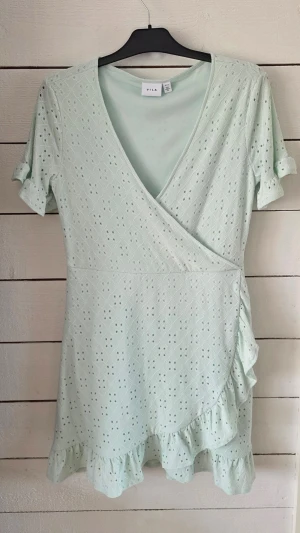 Ljusgrön omlottklänning från Vila - Söt ljusgrön omlottklänning från Vila med korta ärmar, volangdetaljer och broderat hålmönster. Klänningen har v-ringning och är perfekt för varma dagar. Tillverkad i mjukt material som känns skönt mot huden.