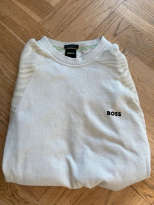 Vit tröja från BOSS med logga - Snygg vit tröja från BOSS med diskret svart logga broderad på bröstet. Tröjan har rund halsringning och ribbade detaljer vid ärmar och hals. Tillverkad i mjukt material som känns skönt mot huden. Perfekt för en clean och stilren look.