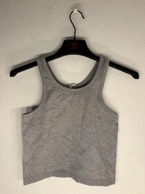 Grå ribbad cropped tanktop - Helt oanvänd. Säljer en enkel och stilren grå ribbad cropped tanktop med breda axelband och rundad halsringning. Perfekt att matcha med jeans eller kjol för en avslappnad look. Materialet är mjukt och stretchigt för extra komfort.