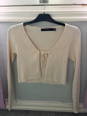 Beige ribbad kofta från Vera Moda - Trendig beige ribbad kofta från Vera Moda med långa ärmar och knytning framtill. Kort, croppad modell med djup urringning och mjukt stretchigt material som passar perfekt till både jeans och kjol.