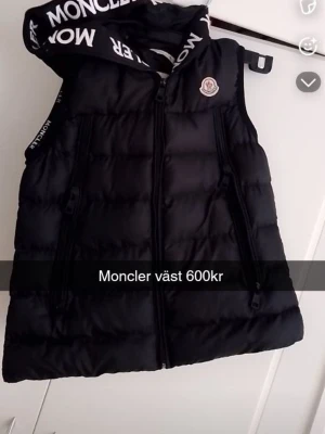 Svart dunväst från Moncler - Snygg svart dunväst från Moncler med huva och dragkedja framtill. Västen har två fickor med dragkedja och Moncler-logga på bröstet. Huva med vit Moncler-text ger en cool kontrast. Perfekt för lager-på-lager och streetwear.