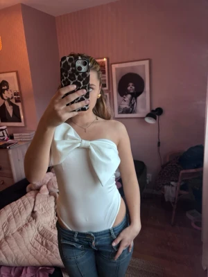 Vit axelbandslös body med rosett - Trendig vit body med stor dekorativ rosett framtill och axelbandslös design. Sitter tight och har en stilren, elegant look. Perfekt att styla med jeans eller kjol för en snygg outfit.