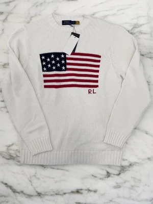 Vit stickad tröja Polo Ralph Lauren - Vit stickad tröja från Polo Ralph Lauren med amerikansk flagga i rött, vitt och blått på bröstet samt RL-broderi. Tröjan har rund halsringning, ribbstickade muddar och är tillverkad i ett mjukt material.