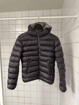 Colmar jacka dam - Snygg svart dunjacka från Moncler med huva och två dragkedjeförsedda fickor. Jackan har quiltad design med breda kanaler och en diskret Moncler-logga på ärmen. Perfekt för kalla vinterdagar och riktigt lätt att bära.