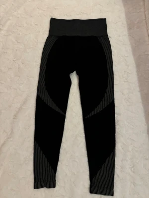 Svarta sportleggings med gråa detaljer - Snygga svarta sportleggings med högt midjeband och gråa ränder längs sidorna och benen. De har en tight passform och är tillverkade i ett stretchigt material som passar perfekt för träning eller gym. Mönstret ger en sportig look och leggingsen är bekväma att röra sig i.