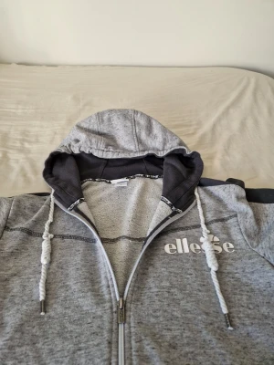 Ellesse zip hoodie - Snygg gråmelerad hoodie från Ellesse med dragkedja hela vägen, vita snören i huvan och svart insida på huvan. Loggan är tryckt i vitt på bröstet. Hoodien har två fickor framtill och en mörkgrå rand längs ena ärmen. Perfekt för chill eller streetwear.