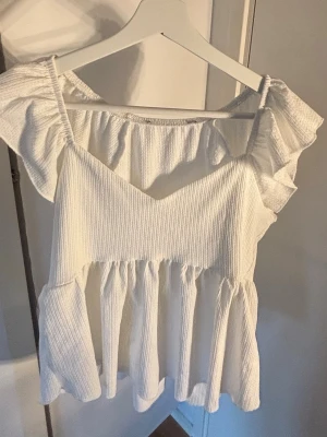 Vit volangblus med v-ringning - Supersöt vit blus med v-ringning och volangdetaljer på axlarna. Blusen har en luftig passform och är tillverkad i ett ribbat material som ger en skön känsla. Perfekt för dig som gillar romantiska och feminina toppar.