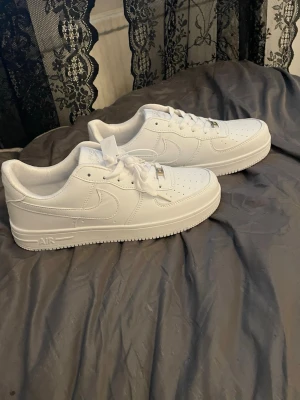 Nike Air Force 1 vita sneakers - Klassiska helvita Nike Air Force 1 sneakers i lågt utförande. Skorna har ovandel i läder, perforerade detaljer på tån och den ikoniska swoosh-loggan på sidan. Tjock sula med AIR-dämpning och metallbricka på snörningen. Perfekta för en clean och stilren look.