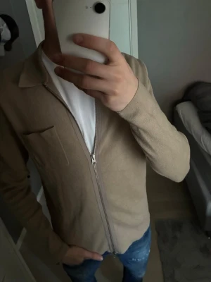 Beige overshirt med dragkedja från Samsøe - Stilren beige overshirt med dragkedja framtill och klassisk krage. Tillverkad i ett mjukt tyg som ger en avslappnad look. Bröstficka på vänster sida och raka ärmar. Perfekt att bära över en t-shirt för en enkel men snygg outfit.