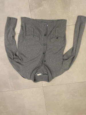 Grå kofta med fickor och knappar - Snygg grå kofta med rund halsringning och knäppning framtill. Två bröstfickor med lock och knappdetaljer ger en cool touch. Ribbstickade muddar vid ärmslut och nederkant. Perfekt att slänga över en t-shirt för en avslappnad look.