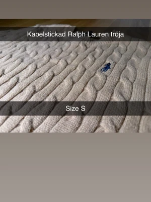 Kabelstickad tröja från Ralph Lauren - Kabelstickad beige tröja från Ralph Lauren med klassisk blå logga broderad på bröstet. Tröjan har rund halsringning och ribbade muddar. Perfekt för dig som gillar tidlös och stilren design.