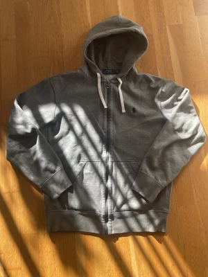 Grå hoodie från Polo Ralph Lauren - Snygg grå hoodie från Polo Ralph Lauren med dragkedja, vit dragsko i huvan och klassisk broderad logga på bröstet. Tillverkad i mjuk bomullsblandning och har känguruficka framtill. Perfekt för en chill och stilren look. Jag vikde in armarna för jag ej hade plats men det är inget fel på dem eller några defekter!